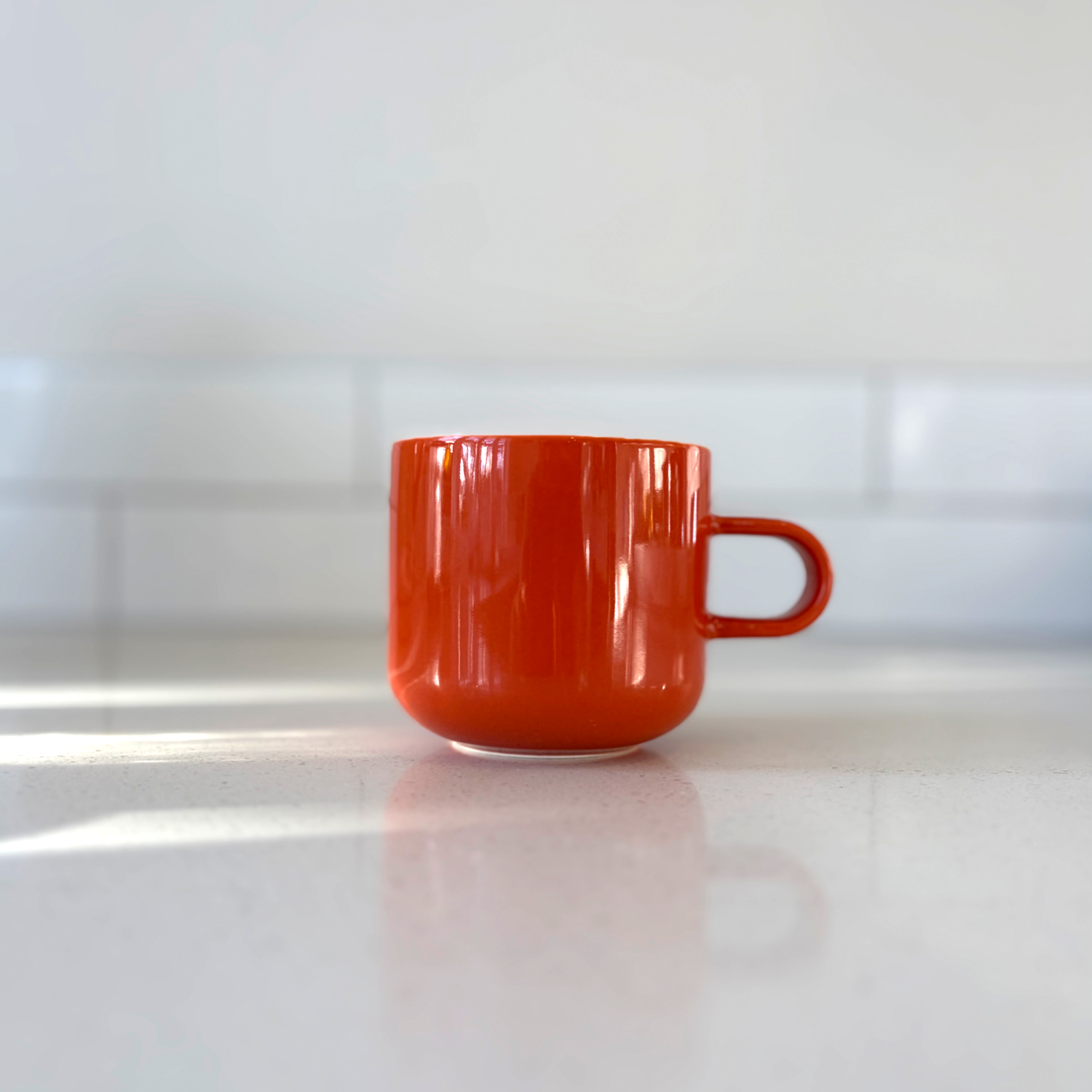 Acme orange ceramic clay mug with a small handle, standing on a light countertop with a tiled backsplash in the background.