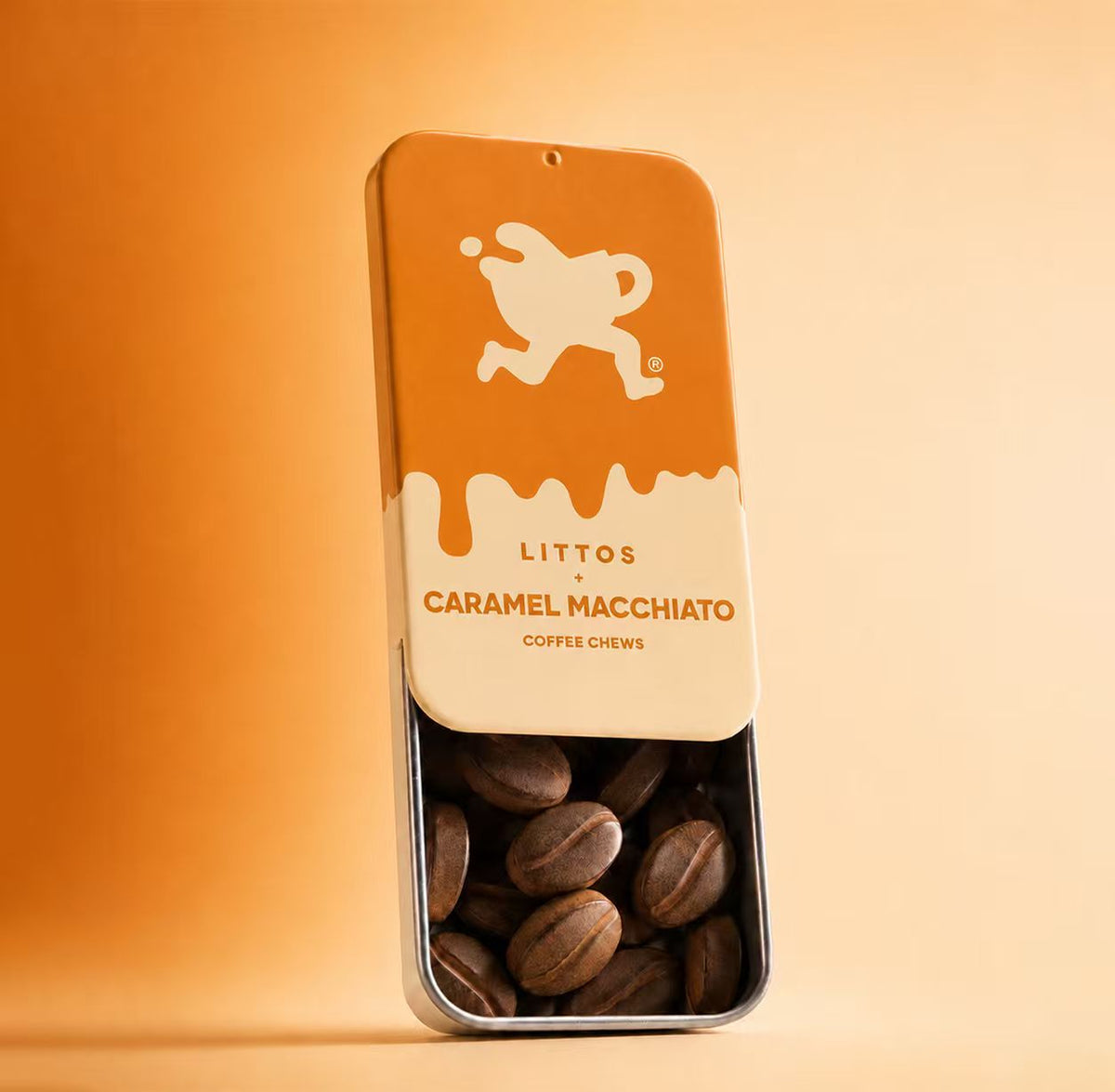 A tall rectangular metal tin of Littos Caramel Macchiato coffee chews stands on a warm beige surface against a matching beige background. The tin has an orange top half with a cream logo, a cream middle section with the product name, and a clear lower half showing brown coffee chews inside.