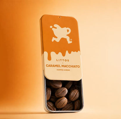 A tall rectangular metal tin of Littos Caramel Macchiato coffee chews stands on a warm beige surface against a matching beige background. The tin has an orange top half with a cream logo, a cream middle section with the product name, and a clear lower half showing brown coffee chews inside.