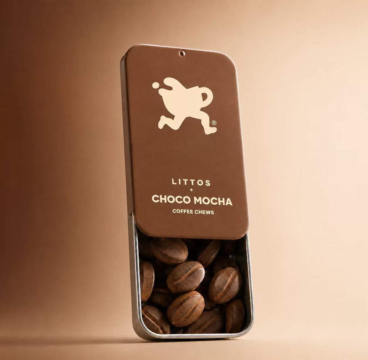 A rectangular metal tin of Littos Choco Mocha coffee chews stands open on a warm beige background. The tin is dark brown with a silver metal rim, and the lower half is pulled open to reveal roasted coffee beans inside.