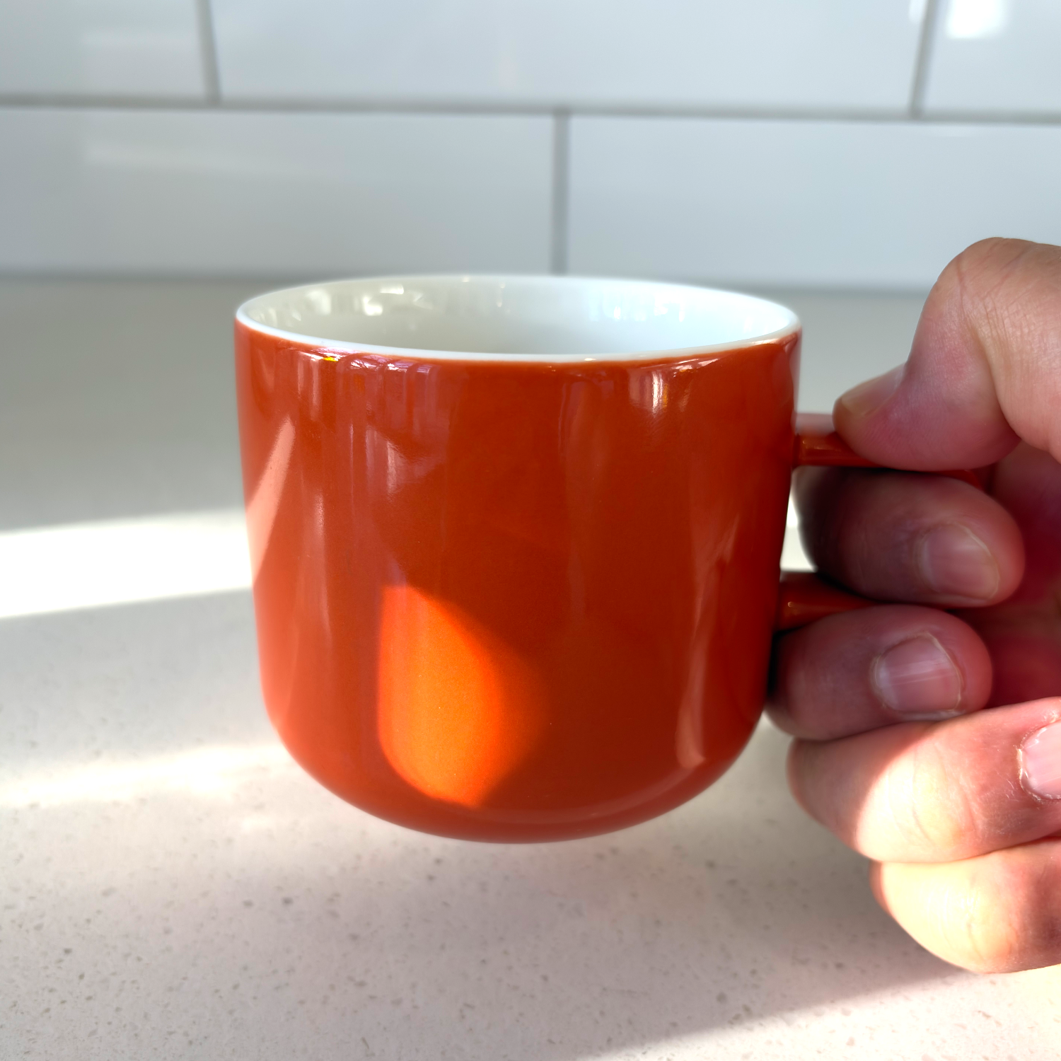 A hand holding acme orange clay ceramic mug with a white interior, set against a white tiled kitchen backsplash and light countertop.