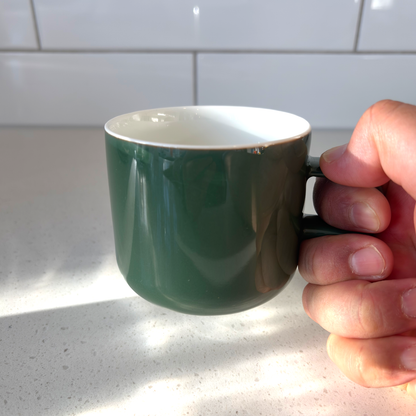 Acme kawakawa green ceramic bobby mug with a white interior, held by a hand against a white tiled kitchen backsplash and light speckled countertop.