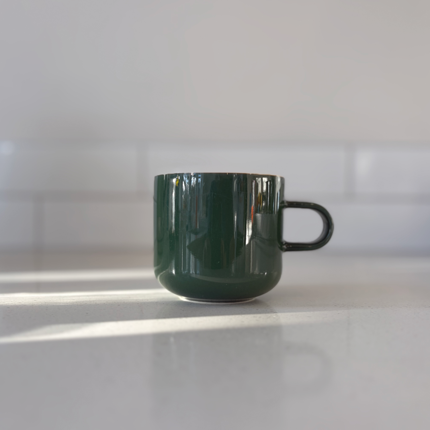 Acme green kawakawa ceramic bobby mug with a glossy finish, sitting on a light gray countertop with soft sunlight and shadows.