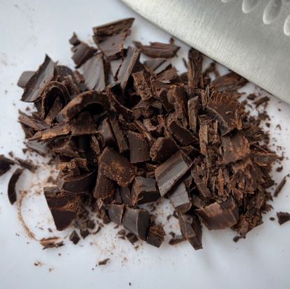 Chopped dark chocolate shavings piled on a white surface with a metal knife blade visible in the top right corner.